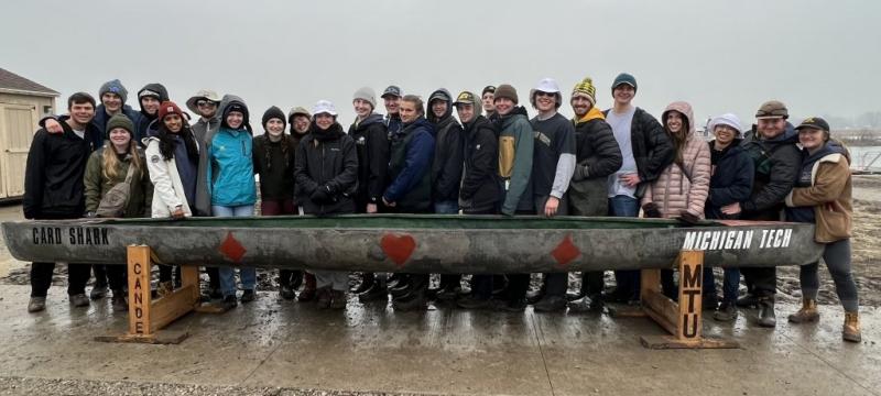 24 members of Michigan Tech Concrete Canoe Team stand behind their canoe Card Shark in a competition at a pond near Wayne State University.