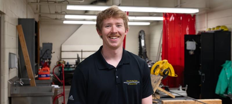 Connor Tokie smiles in the Dow machine shop wearing a black polo that reads â€œSteel Bridge Team at Michigan Techâ€