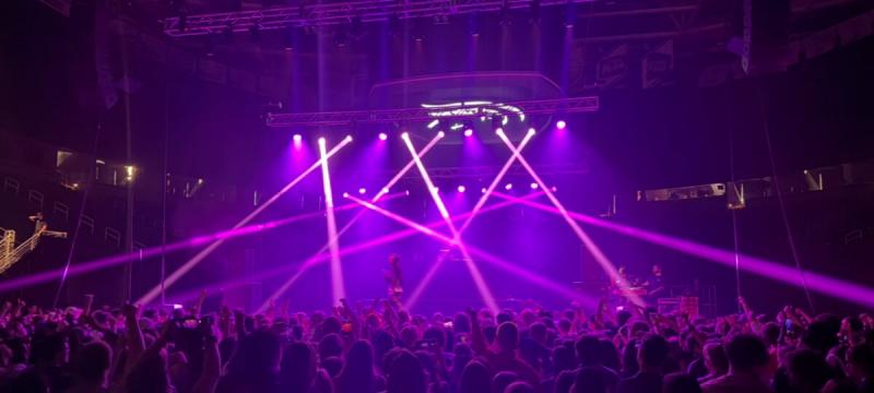 A concert venue filled with people and lit with bright purple beams from multiple stage lights.