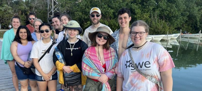 Twelve young people from Michigan Tech smile on a dock with water and small boats behind them as they study abroad in the Yucatan Peninsula.