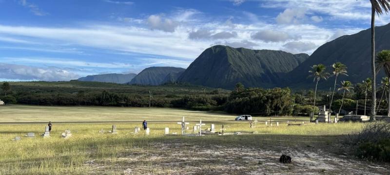 A couple people walking through a field with some grave markers with mountains in the distance.