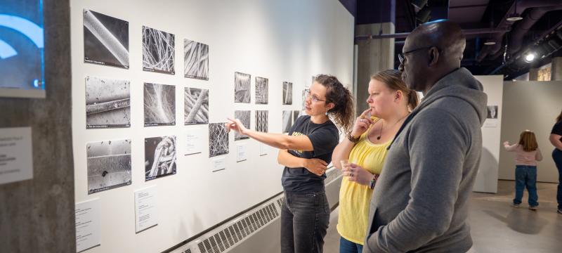 Three gallery attendees observe a collection of nine black and white microscopic images in the Rozsaâ€™s A-Gallery.