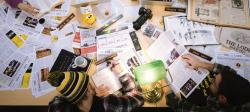 Papers, camera, water bottles clutter a table while students work around it.