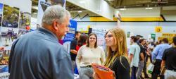 Gray-haired man in blue polo shirt speaks to female student wearing eyeglasses and smiling while holding an orange folder with resumes and employer information in a gymnasium with other students in business attire in background.