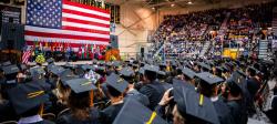 Looking toward the giant American flag behind the stage with many graduate in caps and gowns seated in front and people in the stands at commencement.