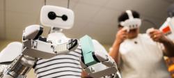 Reachy the Robot grasps two blue blocks as a student in a virtual reality headset controls the robot’s motions.
