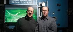Raymond Shaw and Will Cantrell stand facing the camera in front of a green monitor screen mounted to the blue metal cloud chamber behind them.