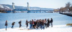 Students carry a concrete canoe from the snowy banks of the Keweenaw Waterway with the Portage Lift Bridge in the background.