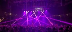A concert venue filled with people and lit with bright purple beams from multiple stage lights.