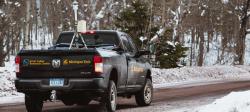 A black truck with the words Great Lakes Research Center and the Michigan Tech logo on the back drives away down a snowy road with an isotopic analyzer mounted in the truck bed.