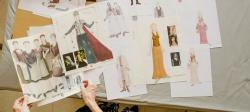 Seven sketches of women in gowns on a table with female hands shown holding them in a costume design shop.