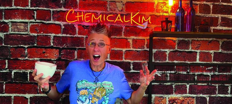 Chemical Kim standing in front of a brick wall with neon Chemical Kim sign holding a bowl and test tube.