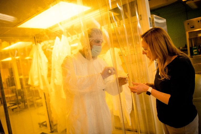 Researcher showing a sample to another person from behind the clean lab barrier.