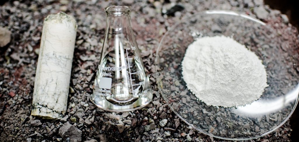 Cored gypsum, a beaker of water, and a plate of plaster of Paris rest on pebbles and dirt.