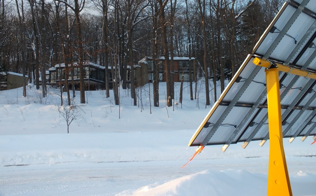 Sunlight shines on solar panels mounted outdoors in the snow in a parking lot near trees