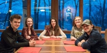 three young women and two young men sitting at a dining room table in a house