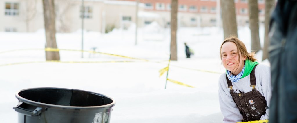 Smiling girl with a black plastic garbage can and snow staked out with a brick building in the background. 