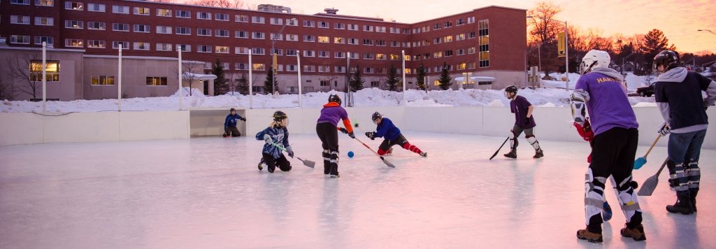 Seven students adorned in helmets knee pads, stand on an ice rink with brooms in hand. 