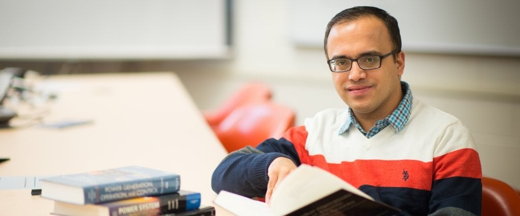 Man with a stack of three books holding a book at a long table with a white screen behind him 