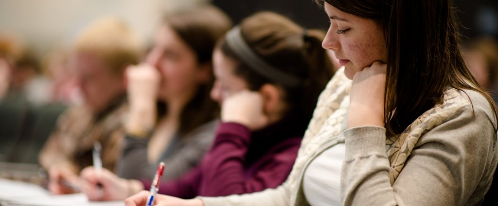 Students sit in a row in a lecture hall and the front student in focus is wearing a tan zip-up sweatshirt.