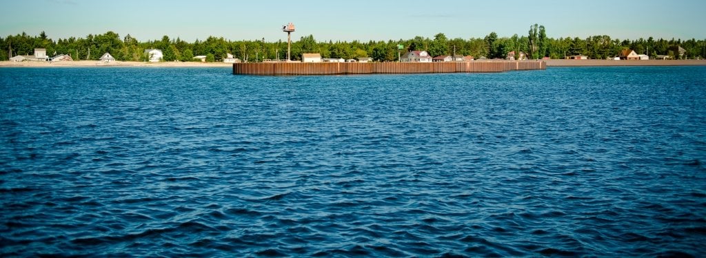 Houses along the shoreline of Grand Traverse Harbor with the white sand beaches on the left side of the harbor mouth breakwall and the gray stamp sands visible on the right side of the breakwater.