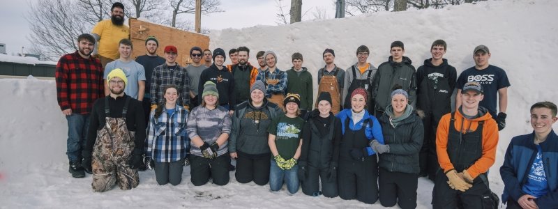 A group of male and female students by a snow wall and wooden form with a male adult