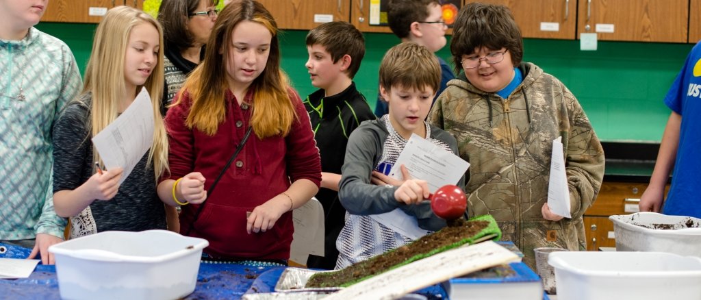 A group of students stand around a table where a pan filled with soil and fake grass sits at a steep angle, one boy in a gray shirt pours water.  