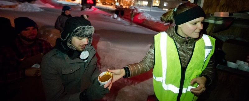 young woman in green vest hands chili cup from a van to a young man in line with other people waiting