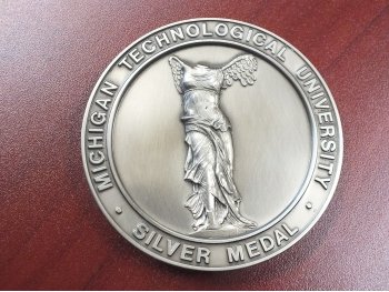 A round silver medal with a woman's torso with wings of coins and Michigan Technological University printed around the rim