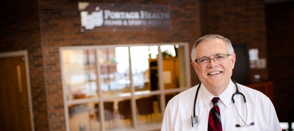 Doctor with stethoscope around his neck in front of brick wall with Portage Health sign on it