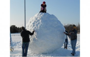 a young woman on top of a giant snowball and other students standing around the giant snowball outdoors in the snow