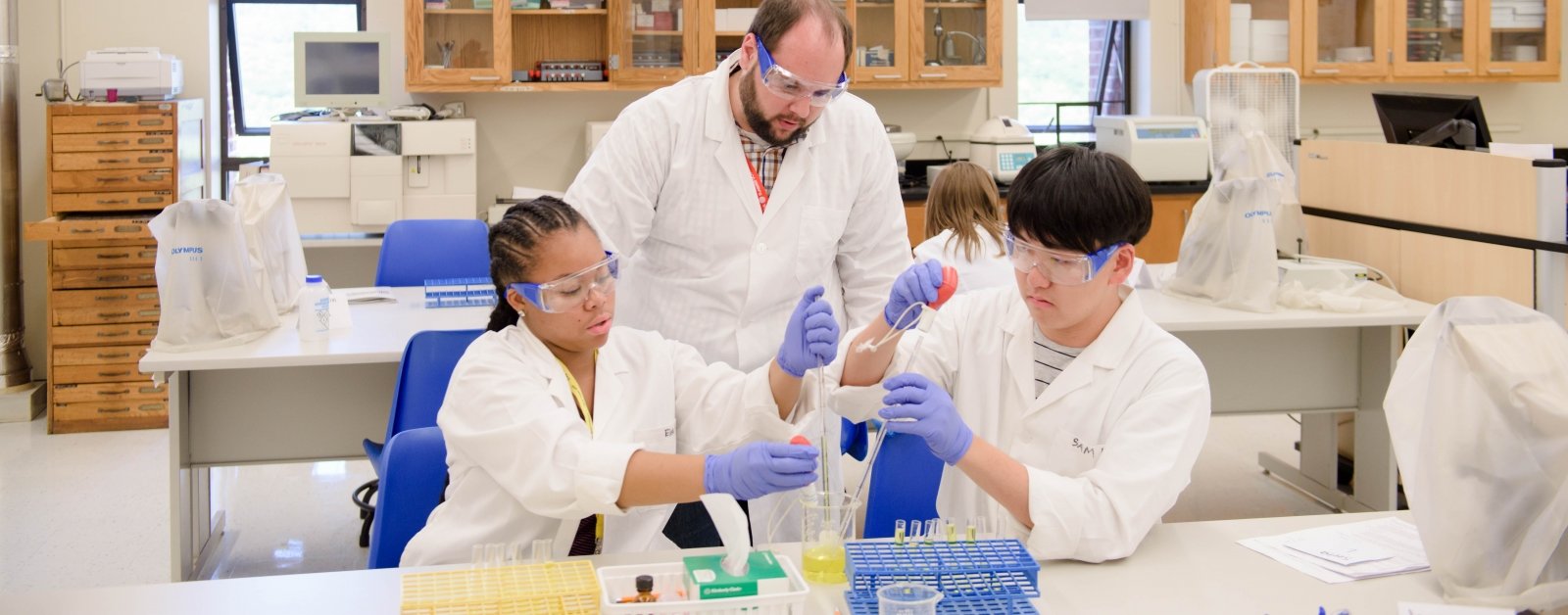 A teacher stands behind two seated students wearing rubber gloves as they drop liquid into a beaker. 