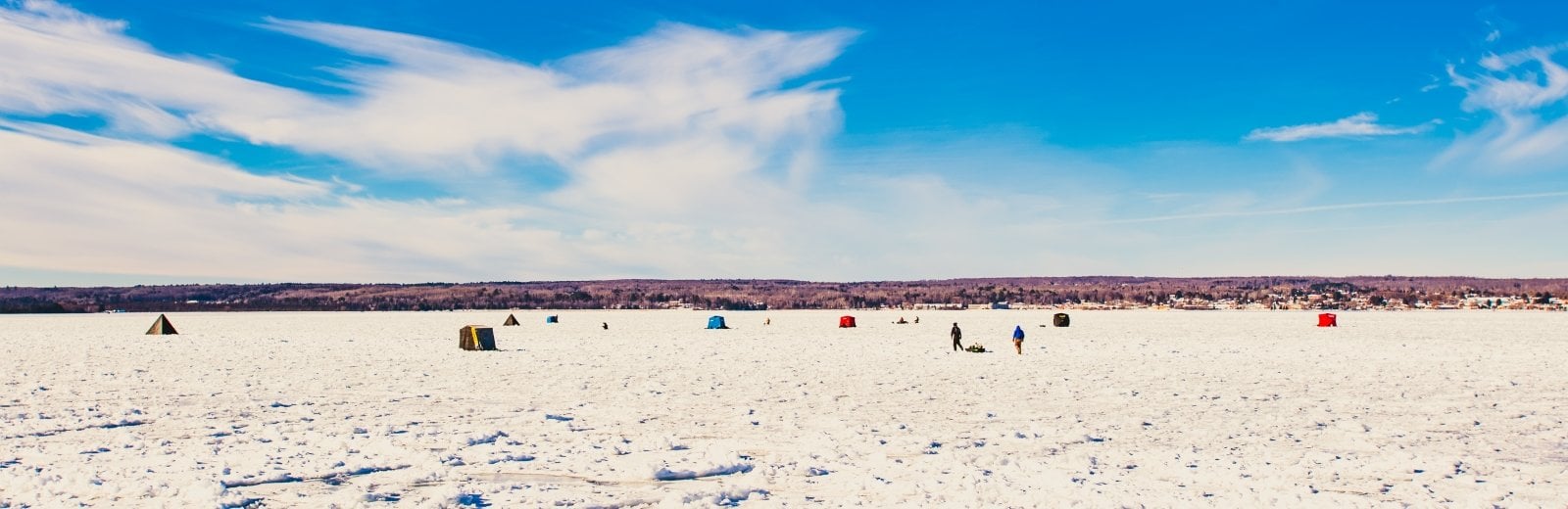 A wide landscape with blue sky and white ice dotted with people and ice fishing huts. 