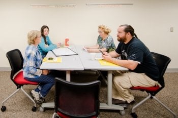 Four people sit around a table taking notes with an Objibwa language banner hanging behind them.