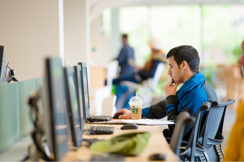 A student sits in front of a computer monitor at a table with several computers in a row.