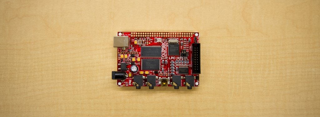 A red circuit board sits on a blond wood table.
