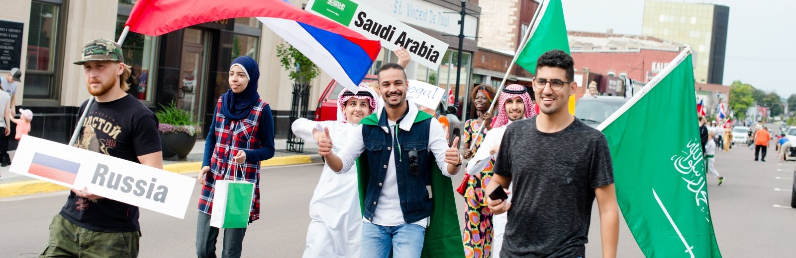 men carrying Saudi Arabia, Senegal, Russia flags and a woman carrying a bag smiling in a parade on a downtown street in a small town 