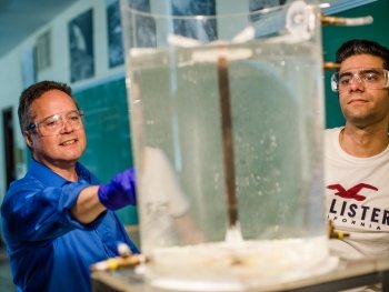 Two men look at water in a tabletop plastic tank with a PVC-pipe sprinkler attached.