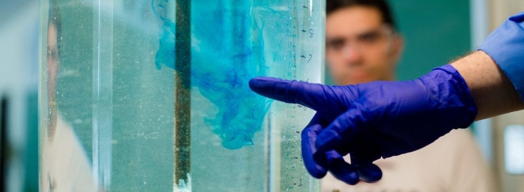 A man's gloved hand points to colorant swirling in a water tank.