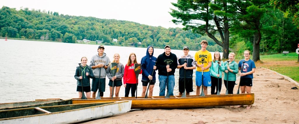 11 young men and women with canoe paddles and two canoes stand on the beach by the water