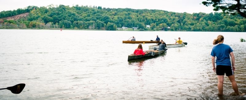 students in shorts on the water by a beach paddling in two canoes