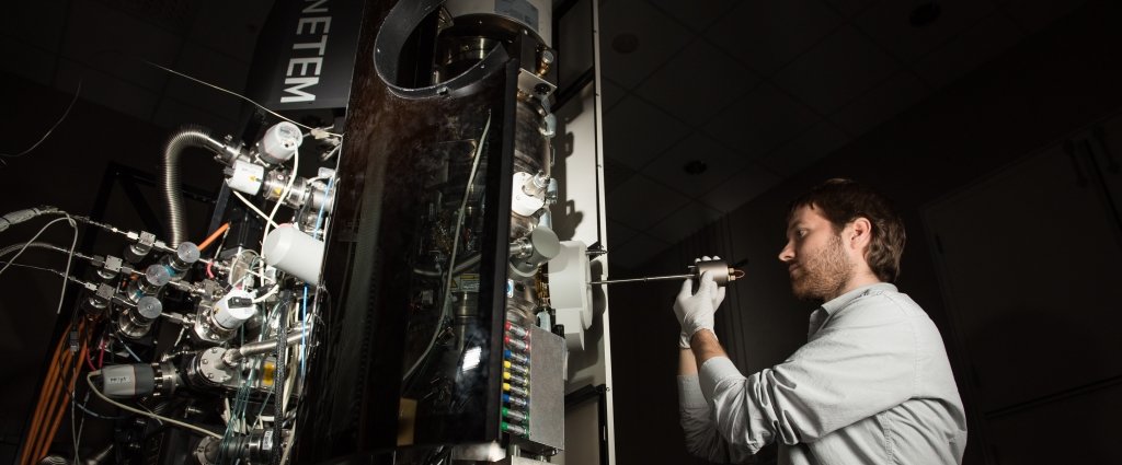 A man holding a screwdriver-sized instrument stands next to a tall machine with exposed wires and circuits.