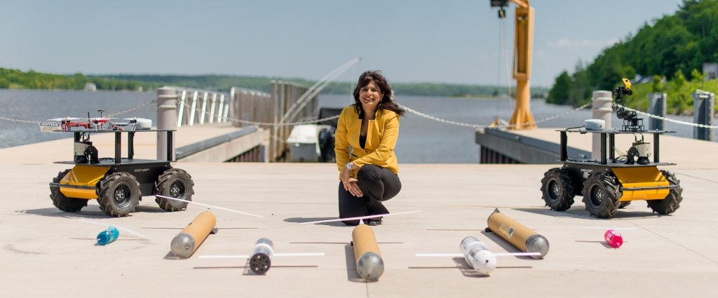 A woman kneels on a boat deck surrounded by a semicircle of different robot prototypes.