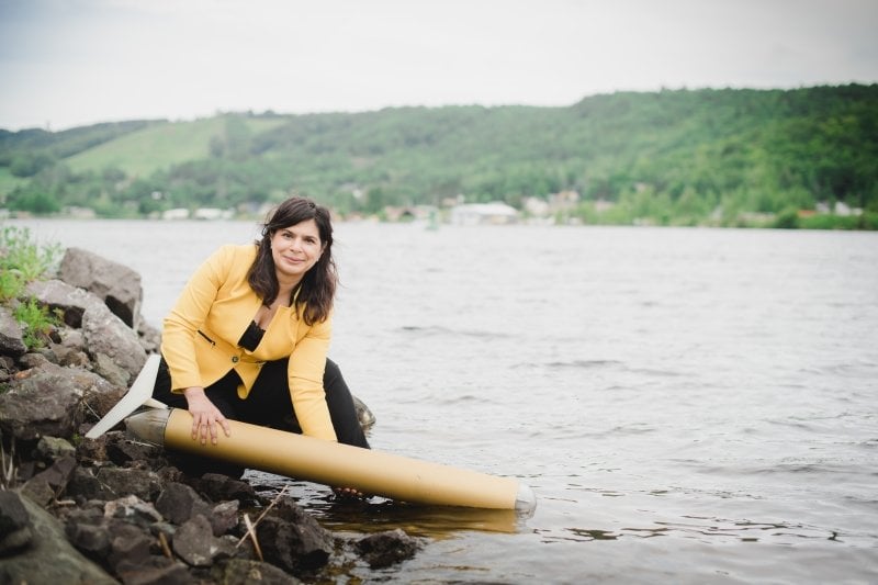 A woman kneels on a rocky shore holding a tan-colored autonomous underwater vehicle