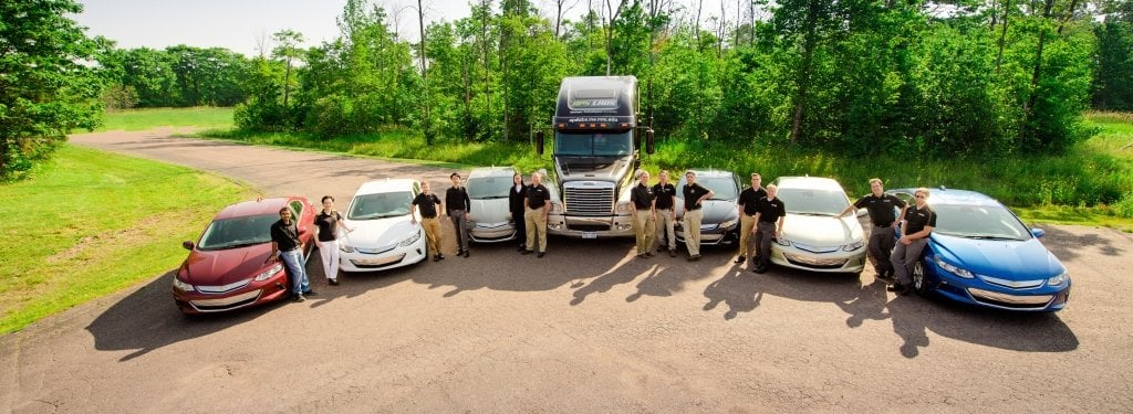 People in black polo shirts stand in between a semicircle of cars and one semi-truck. 