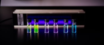 Six small bottles on a laboratory counter filled with multi-colored fluorescent dyes.