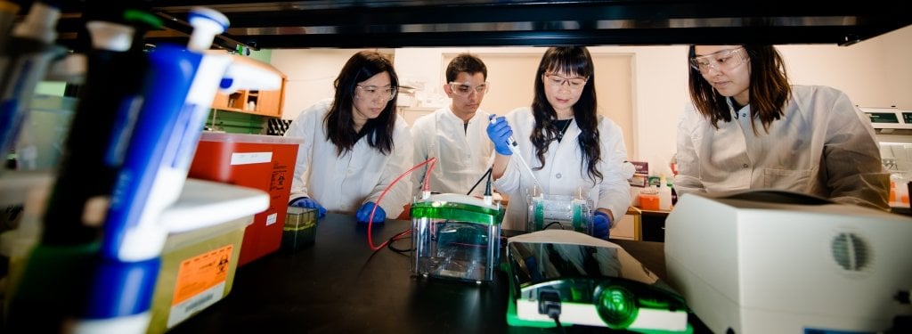 Four people in lab coats watch a female scientist use a pipette to suck liquid from a beaker.