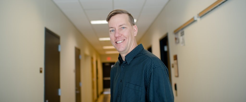 A man wearing a blue shirt smiling in a door-lined hallway 