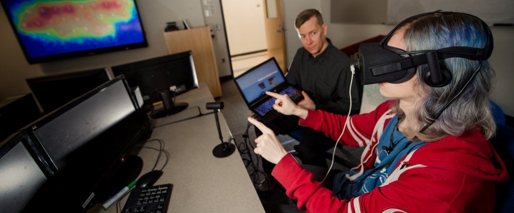 a male and a female sit in front of a computer the man is holding a laptop and the female is wearing a virtual reality headset and typing in space on an envisioned keyboard in a computer science classroom