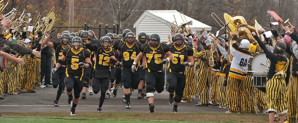 a football team running onto a grassy field in an outdoor stadium with pep band members in striped clothes and funny hats cheering for them